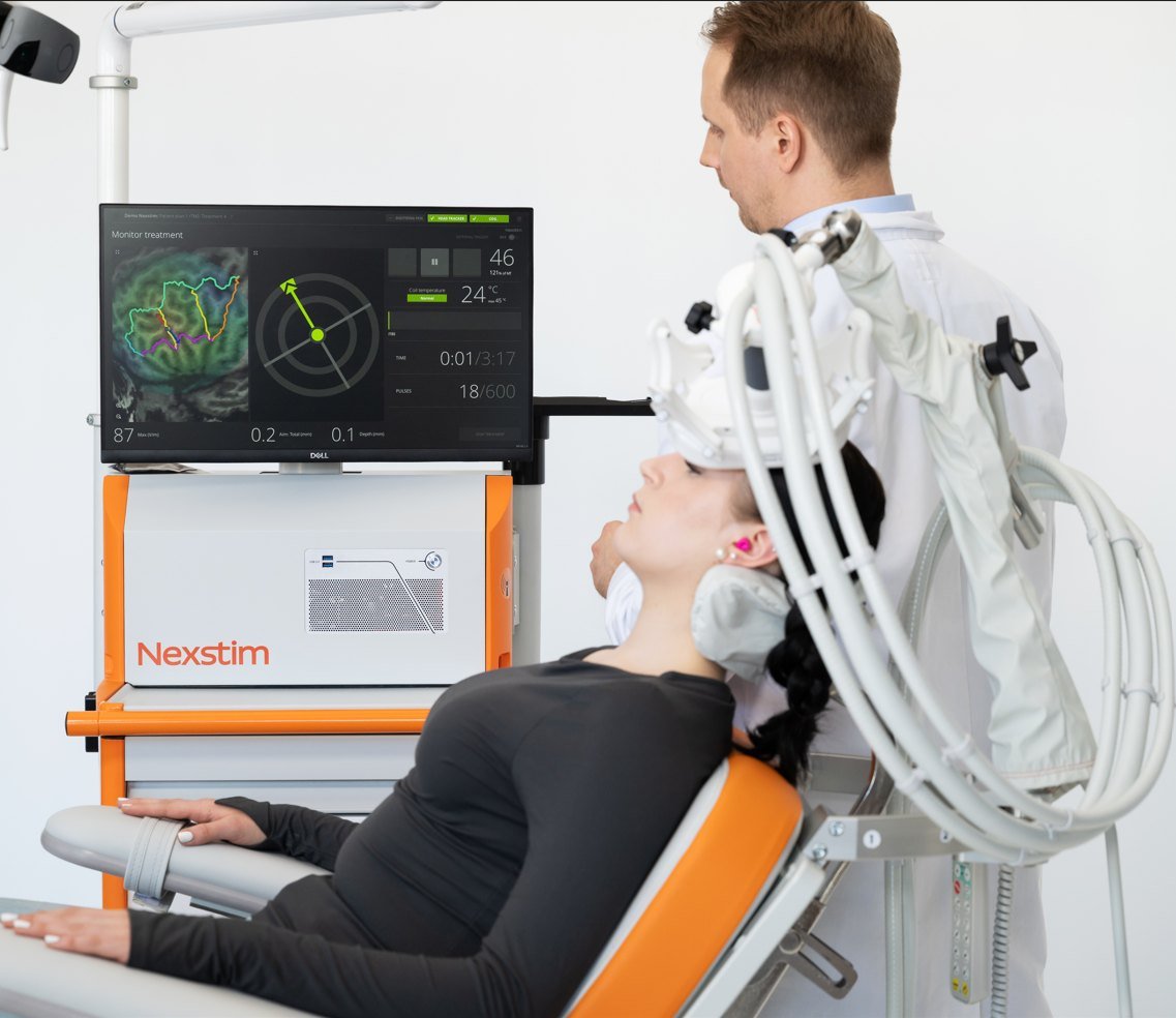 A patient rests comfortably in the treatment chair beside the Nexstim NBS system while a clinician monitors the navigation display during a session.